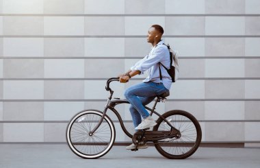 Şehir merkezindeki tuğla duvarın yanında bisiklet süren kulaklıklı zenci adamın yan görüntüsü.