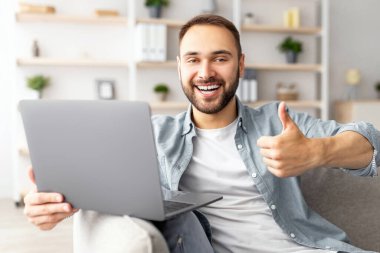 Evde dizüstü bilgisayar kullanan heyecanlı bir milenyum adamı, el hareketini gösteriyor, online çalışmayı ya da çalışmayı öneriyor. Genç erkek serbest çalışan internet üzerinden iletişim kuruyor, iş toplantısına katılıyor.