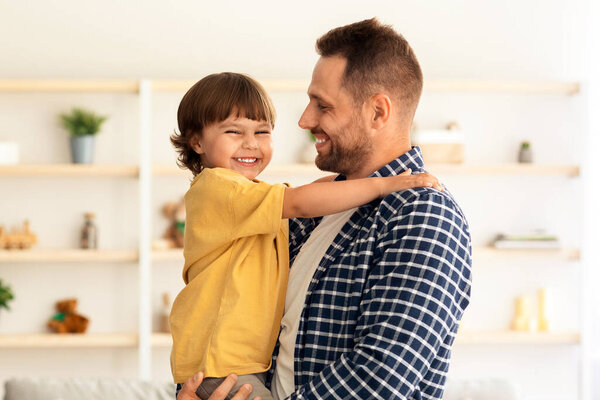 Happy childhood. Cute little boy embracing his loving handsome daddy and smiling to camera, posing together at home interior