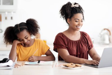 Mutlu Afro-Amerikan liseli kız ödev yapıyor, neşeli çalışan annesinin yanında, mutfak içinde oturuyor. Evde çalışan siyahi bir kadın, dizüstü bilgisayar kullanıyor.