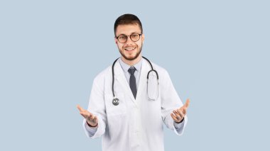 Beyaz üniformalı neşeli erkek doktor. Kamera önünde el hareketleriyle konuşuyor. Mavi stüdyonun arkasında duruyor. Tıp kariyeri, doktor ve hemşire mesleği. Panorama