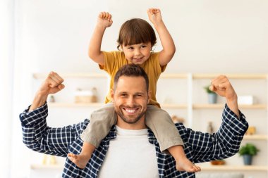 Aile gücü. Küçük oğlunu omuzlarında taşıyan, bisepslerini gösteren ve kameraya gülümseyen genç ve güçlü bir adam. Spor severler konsepti.