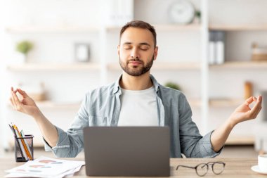 İş yeri stres yönetimi. Sakin Kafkasyalı adam ofisteki bilgisayarın önünde gözleri kapalı meditasyon yapıyor. Genç erkek serbest çalışan, kendini huzurlu ve dengeli hissediyor, masasında yoga yapıyor.