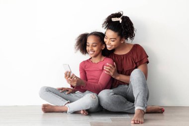 Güzel siyah anne ve kız birlikte cep telefonu kullanıyor, yerde oturuyor, sosyal medya için selfie çekiyor, evde birlikte vakit geçiriyorlar, beyaz boş duvar arka planı, kopyalama alanı.