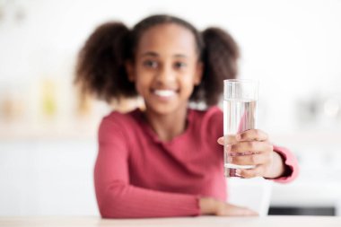 Neşeli Afro-Amerikalı güzel kız masada otururken bir bardak su tutuyor, kameraya gülümsüyor, mutfak içi, kopyalama alanı. Susuzluk, çocuk konsepti için içme suyu.