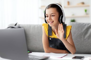 Mesafe öğrenme. Kulaklıklı neşeli bir genç kız, web kamerasına el sallıyor, oturma odasında bilgisayar kullanarak sanal ders alıyor, online eğitimin tadını çıkartıyor.