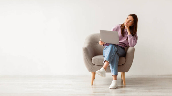 Deadline stress concept. Frustrated young woman sitting in armchair with laptop computer, looking away deep in thought, feeling tired after long working day against white studio wall, full length