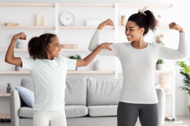 Güçlü Afro-Amerikan anne ve genç kızı spor kıyafetleriyle evde sabah egzersizlerini yaparken kaslarını gösteriyorlar, atletik siyah aile pazılarını gösteriyor ve gülümsüyor.