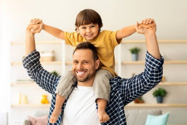Birlikte iyi vakit geçirin. Neşeli genç baba, küçük tatlı oğlunu omuzlarında taşıyor, evde birlikte eğleniyor, kameraya gülümsüyor, boş bir alanda.