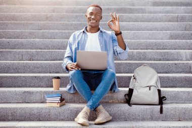 Çevrimiçi eğitim saçmalıkları. Laptoplu, sırt çantalı ve kitaplı Afro-Amerikan öğrenci merdivenlerde iyi bir jest gösteriyor. Gülümseyen siyah adam onaylıyor, e-öğrenmekten mutlu.