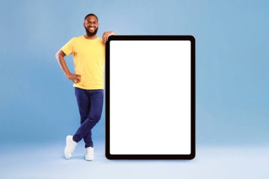 Dev tablete yaslanmış neşeli Afro-Amerikan bir gencin tam boy portresi, mavi arka planda boş ekran gösteriyor, online reklam için boş alan, en yeni uygulama için model