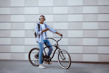 Mobil cihazlı ve kulaklıklı siyahi erkek hippi şehir sokağındaki tuğla duvarın yanında bisiklet sürüyor.