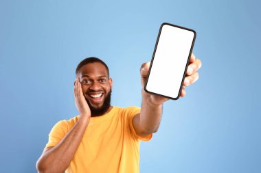 Şu havalı mobil uygulamayı kontrol et. Heyecanlı Afrikalı Amerikalı adam akıllı telefonu boş ekranla sunuyor, web sitesi ya da mavi stüdyo arka planında reklam öneriyor, maket için yer var. Seçici odak