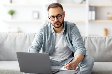 Ciddi bir genç adam dizüstü bilgisayar kullanıyor, internette çalışıyor, iş görüşmesinde ev ofisinden notlar alıyor. Milenyum adamı uzaktan çalışıyor, bilgi yazıyor. Modern teknoloji kavramı