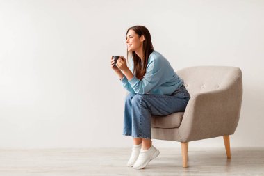 Beyaz stüdyo duvarına karşı rahat bir koltukta kahve molası veren genç bir kadın. Boş alana sahip pankart tasarımı. Büyüleyici milenyum hanımı dinleniyor, sıcak içkinin tadını çıkarıyor.