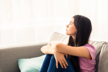 Gençlik depresyonu konsepti. Stresli Hintli genç kız evdeki kanepede oturuyor, pencereden dışarı bakıyor, iletişim eksikliği çekiyor, kendini üzgün ve yalnız hissediyor.