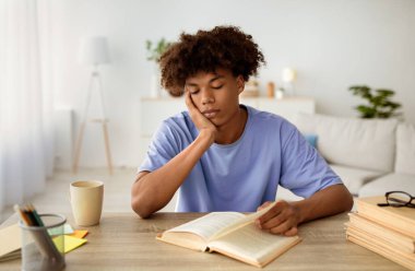 Masasında oturup kitap okuyan, evde ders çalışan sıkıcı siyahi bir genç. Afrikalı Amerikan gençliği uzaktan öğreniyor, ödev yapıyor, sınava hazırlanmak için ders kitabını kullanıyor, kapalı alanda.