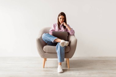 Dizüstü bilgisayarlı koltukta oturan ciddi genç bayan uzaktan çalışıyor ya da beyaz stüdyo duvarına karşı video konferansı yapıyor. Milenyum kadını internet üzerinden Webinar izliyor.