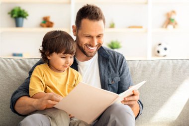 Bebeklerle hafta sonu aktiviteleri. Neşeli taşıyıcı baba küçük oğluna kitap okuyor, bebek baba dizlerinin üzerinde peri masalının tadını çıkarıyor, evde kanepede oturuyor.