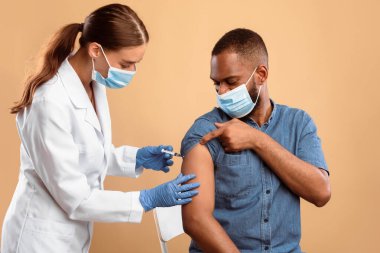 Grip salgını sırasında aşı ve sağlık hizmetleri. Yüz maskeli genç bir doktor bej rengi stüdyo geçmişi olan Afrikalı Amerikalı hastaya grip aşısı yapıyor. Yapay dokunulmazlık kavramı