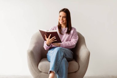 Evdeki gibi rahat ol. En sevdiği kitabı okuyan neşeli genç bir kadın beyaz stüdyo arka planında koltukta oturuyor. Güzel bir milenyum bayanı haftasonu ilginç bir hikayenin tadını çıkarıyor.