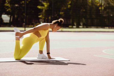 Açık hava eğitim kavramı. Kendine güvenen sportif siyah kadın bacak kaslarını ve baldırlarını yoga minderinde geriyor, sahada egzersiz yapıyor, kulaklık takan, sarı spor giyimli ve beyaz spor ayakkabılı kadınlara odaklanıyor.