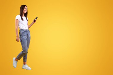 İletişim, Teknoloji ve Yaşam Tarzı Konsepti. Cep telefonu kullanan, turuncu stüdyo arka planında izole edilmiş yürüyen, gülümseyen genç bayanın tüm vücut uzunluğu, panoramik afiş, serbest alan