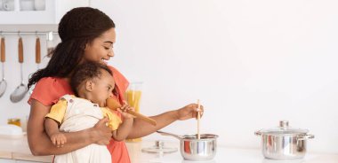 Güzel anne mutfakta yemek pişiriyor, küçük oğlu kucağında, mutlu Afrikalı Amerikalı kadın ev işi yapıyor ve bebek bebekle ilgileniyor, aile için sağlıklı yemekler hazırlıyor,