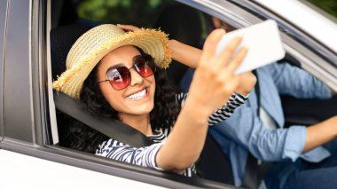 Olumlu genç bir kadın cep telefonuyla selfie çekerken sevgilisiyle araba gezisine çıkıyor sosyal medyada arkadaşlarıyla mutlu fotoğraflar paylaşıyor yaz şapkası ve güneş gözlüğü takıyor, seyahat konseptini kullanıyor.