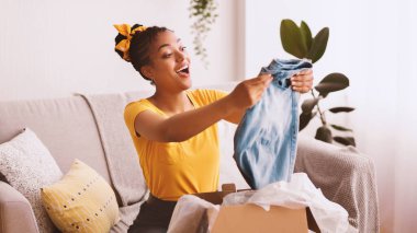 Teslimat Konsepti. Gülümseyen siyah kadın yeni kot pantolon tutuyor, karton paketleri açıyor, evdeki kanepede oturuyor, boş yer var. Alışverişten ve kıyafet sevkiyatından memnun olan heyecanlı bayan.