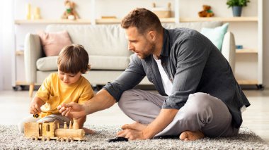 Çocuk gelişimi. Genç ve mutlu bekar baba, tatlı oğluyla evde vakit geçiriyor, yerde oturuyor ve ahşap tren oynuyor, boş yer var.