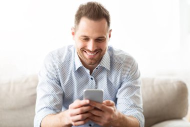 Sosyal ağda mesajlaşma. Modern akıllı telefon cihazı kullanan, evdeki koltukta oturan mutlu bir milenyum adamı.
