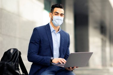 Arap girişimci koruyucu yüz maskesi takmış iş merkezinin yanındaki sokakta oturuyor, laptop kullanıyor, iş sunumundan önce konuşmayı kontrol ediyor, kopyalama alanını kopyalıyor. COVID-19 salgını varken iş dünyası
