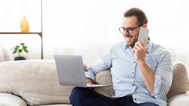 Serbest Kavram. Gözlüklü tatmin olmuş erkek yönetici telefon görüşmesi yapıyor, dizüstü bilgisayar tutuyor, oturma odasında kanepede oturuyor.