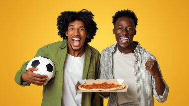 İki heyecanlı siyahi erkek futbol topu ve İtalyan pizzasıyla boks yapıyor, Afro-Amerikan futbol fanatikleri bej arka planda birlikte eğleniyor, Neşeli erkek arkadaşlar favori takımı destekliyor, bedava uzay.