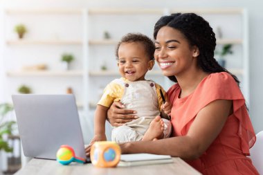 Genç ve mutlu bir anne dizüstü bilgisayarda çalışıyor ve evde bebeğe bakıyor, Afrikalı bir bayanın kucağında sevimli bir çocuk, bilgisayarında daktilo yazıyor, doğum izninde uzaktan kumanda işini seviyor, boş bir alanda çalışıyor.