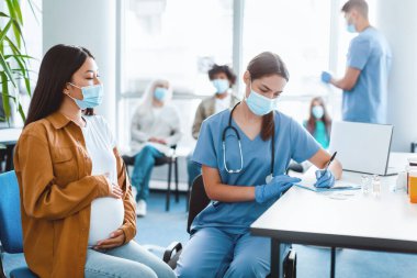 Grip aşısı konsepti. Maskeli Kadın, Coronavirus aşısından önce hamile kadın hastayla konuşurken notlar alıyor. Hastane odasında oturuyor, Asyalı genç bayan göbeğine sarılıyor.