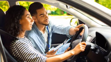 Yakışıklı Arap sürücü eğitmeni elinde direksiyon, yolda nasıl araba süreceğini gösteriyor, esmer kadın sürücü kursunda ders alıyor, yan görüş, yakın çekim.