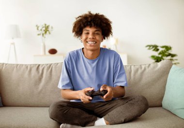 Ev eğlenceleri. joystick 'li mutlu siyah bir genç kanepede bilgisayar oyunu oynuyor, tam boy. Video oyunlarıyla uğraşan Afrikalı Amerikan gençliği