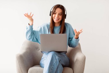Kulaklıklı mutlu bir beyaz kadın dizüstü bilgisayarında iş görüşmesi yapıyor, beyaz stüdyo arka planında rahat bir koltukta oturuyor. Webinar 'a katılan Milenyum öğrencisi uzaktan iletişim kuruyor.