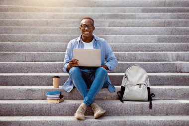 Çevrimiçi eğitim kavramı. Dizüstü bilgisayarı ve kitap yığınıyla şehir merkezindeki merdivenlerde oturan zeki siyahi adam.