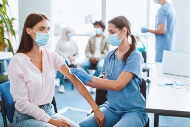 Toplu Aşı Sağlık Merkezi, Gobal Bağışıklık Kampanyası. Tıbbi maskeli kadın doktor ve eldivenler genç bayana grip aşısı enjekte etmeye hazırlanıyor, koluna pamuk pedi sürüyor.