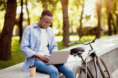Mesafe çalışma konsepti. Bisikletli neşeli Afro-Amerikalı adam şehir parkında dizüstü bilgisayarla çalışıyor, boş bir alanda. Millennial Black Freelancer dışarıda proje yapıyor.
