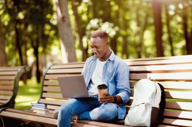 Uzaktan eğitim konsepti. Sırt çantası, kitap ve dizüstü bilgisayarı olan havalı siyah öğrenci parkta online derse katılıyor. Milenyum Afrikalı Amerikalı adam dışarıda web konferansına katılıyor.