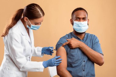 Koruyucu maskeli Afro-Amerikalı adam bej renkli stüdyo arka planında kadın doktordan grip aşısı alıyor. Genç hemşire, aşılayan adam hasta. Dokunulmazlık kampanyası
