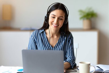 Kulaklıklı kadın, modern ofisteki dizüstü bilgisayarın başına oturmuş video görüşmesi yapıyor. Uzak İş İletişimi Konsepti.