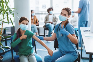 Grip aşısı. Aşılanmış ergen kız ve hemşire klinikte koruyucu maske takarak başparmak hareketi yapıyorlar. Kadın Doktor ve Küçük Hasta Bağışıklığı Onaylıyor