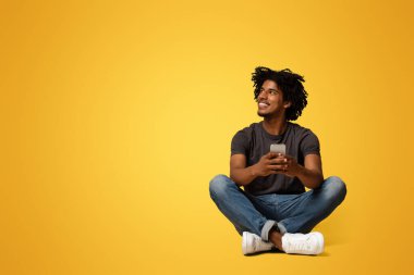 Mobil Teklif. Elinde akıllı telefonuyla gülümseyen genç bir zenci bej renkli stüdyo arkaplanındaki fotokopi odasına bakıyor, iletişim için modern aletler kullanmaktan hoşlanıyor, panorama