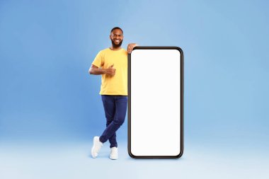 Yeni mobil uygulama. Yakışıklı Afro-Amerikan genç adam büyük ekranlı cep telefonunun yanında duruyor, başparmağını kaldırıyor, havalı bir web sitesi tavsiye ediyor, mavi üzerine reklam için modelleme öneriyor.