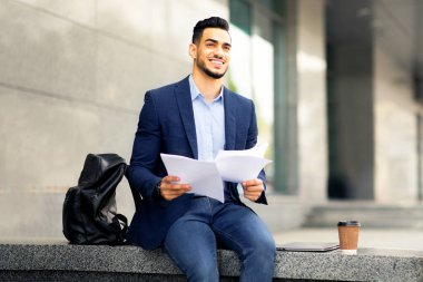 Neşeli genç ortadoğulu girişimci sunumdan önce hazırlanıyor, ofis binasında sırt çantasıyla oturuyor, gazete tutuyor ve kahve içiyor, fotokopi çekiyor.